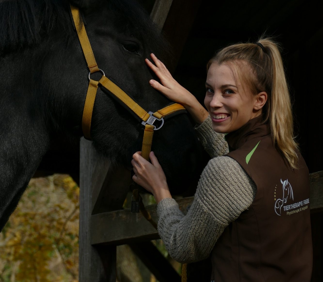 Junge Frau lächelt und streichelt ein schwarzes Pferd unter einem Dach.